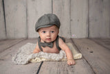 A newborn 3 month old infant wearing a gray herringbone classic vintage style cabbie hat, bowtie, and shorties suspenders set photo prop lays on his belly on a soft blanket, looking at the camera. Rustic wood-look backdrop coordinates with the gray newsie outfit and blanket.